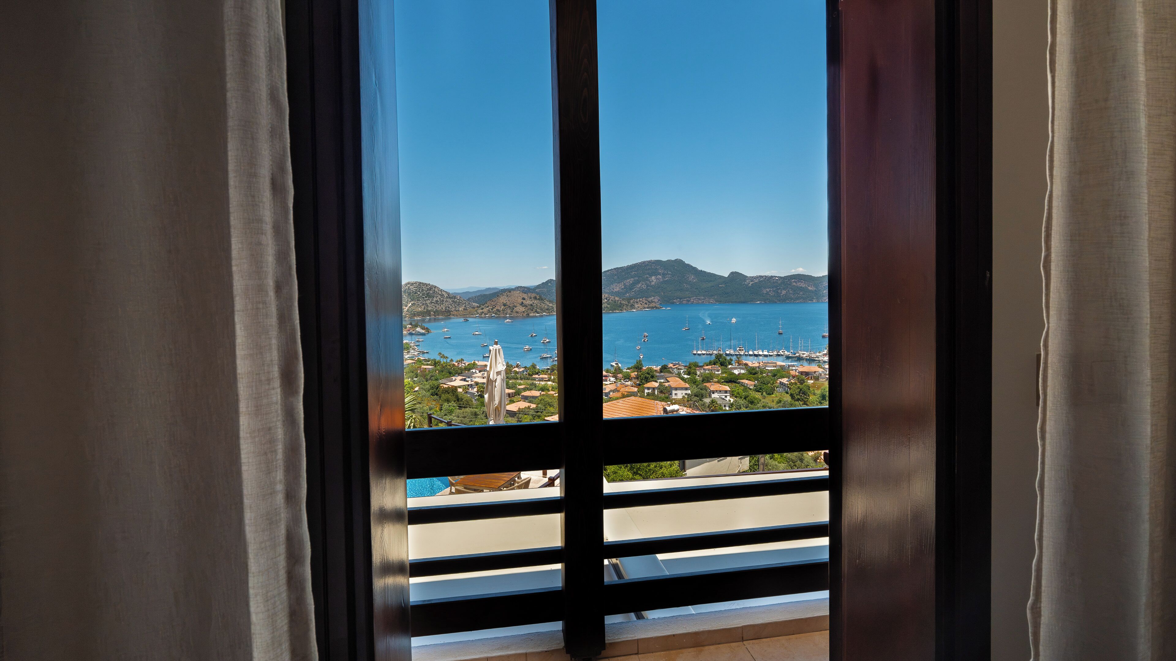 A view through a window with curtains, revealing a coastal landscape with boats, houses, and mountains.