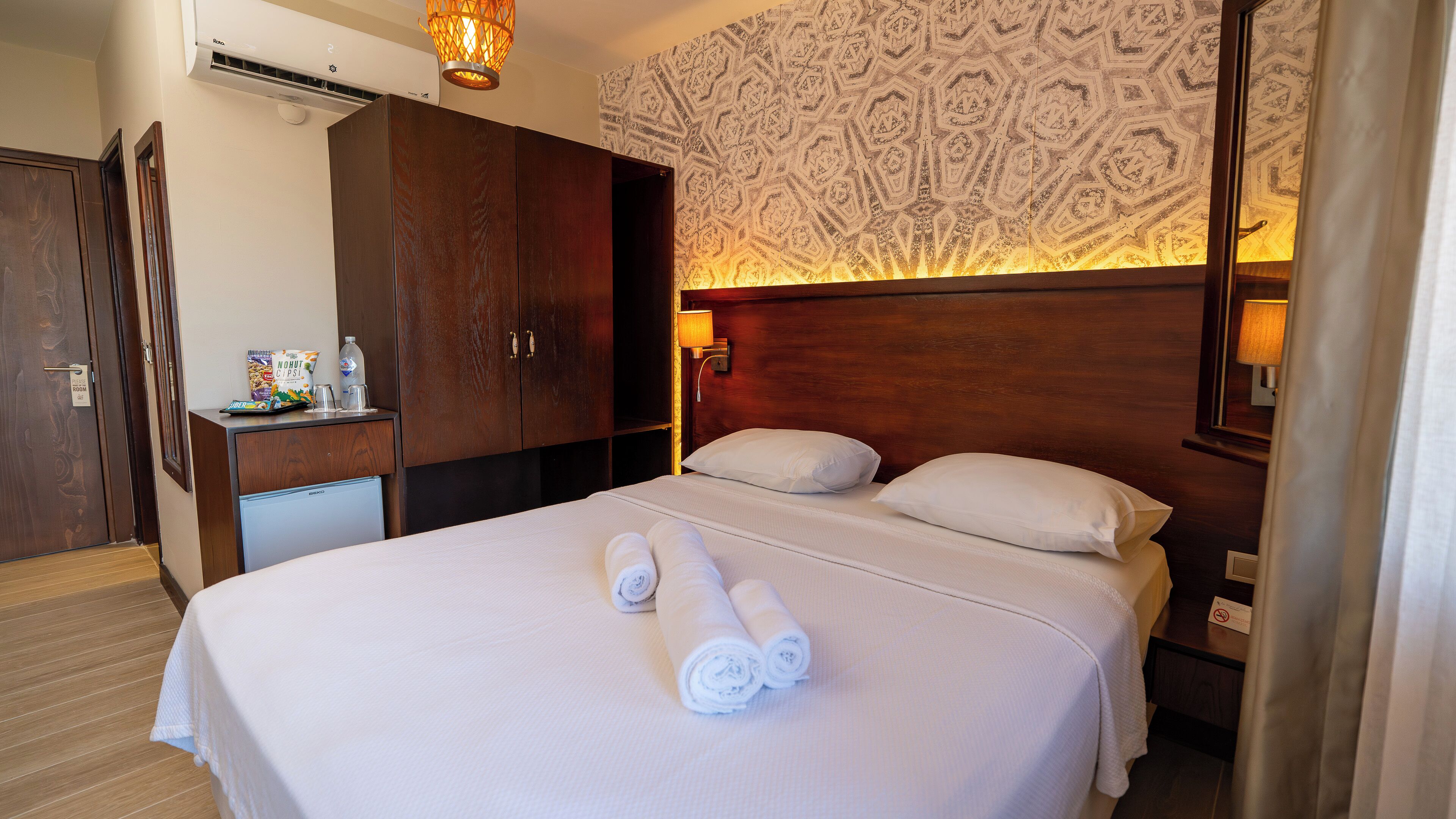 Une chambre d’hôtel avec un lit, deux oreillers, une table de chevet, un bureau avec une lampe, un minibar et un mur décoré d’un motif.