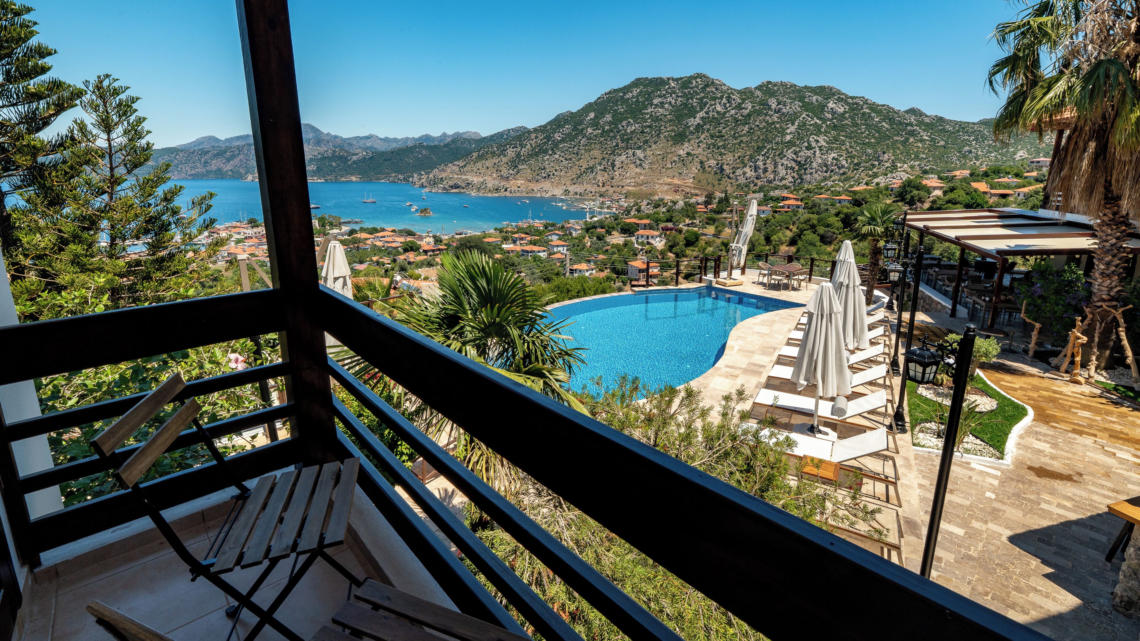 A pool area with lounge chairs and umbrellas, overlooking a coastal town and mountains.