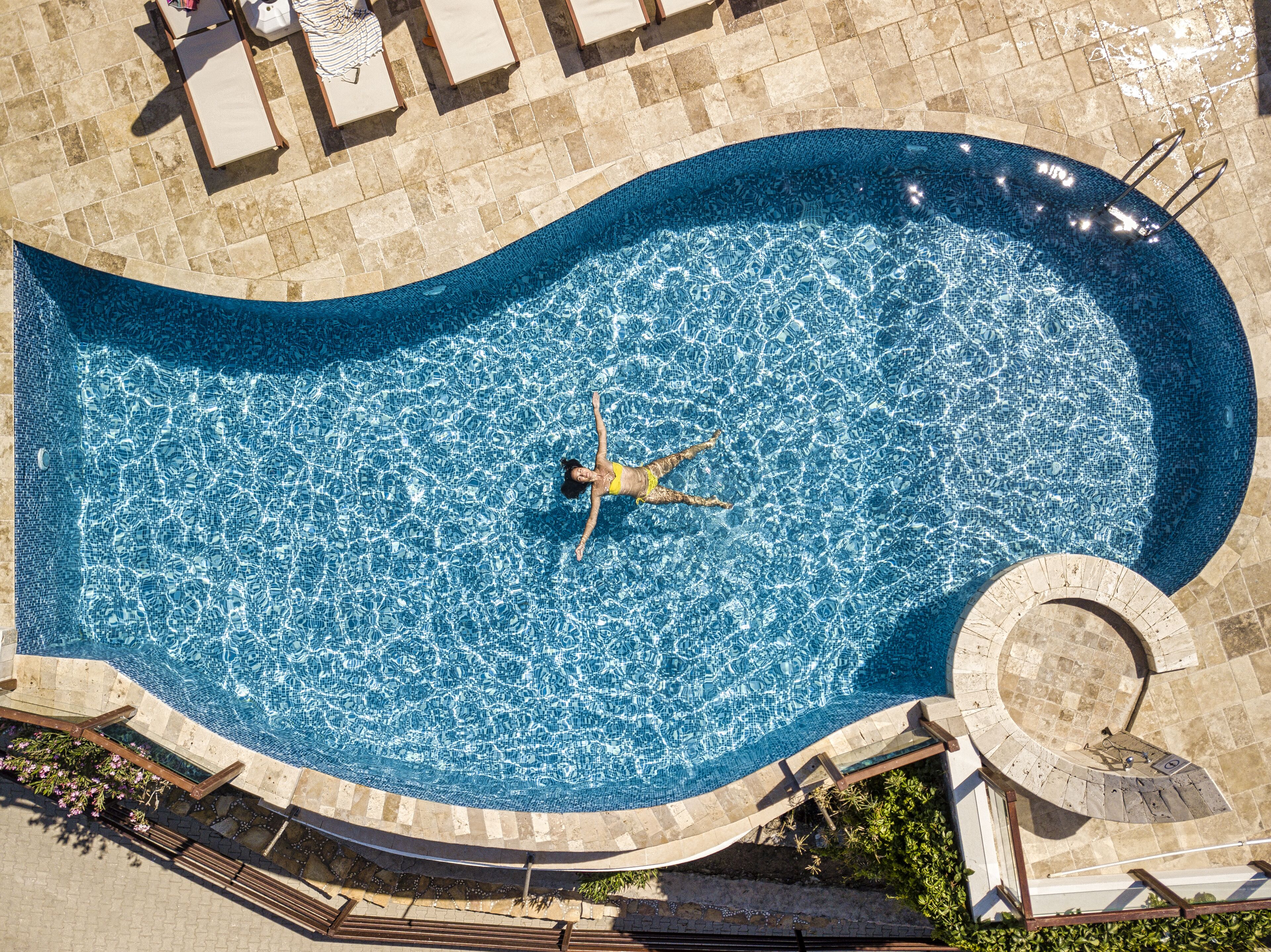 Vista aérea de una piscina con una forma curva distintiva, rodeada por un deck de baldosas y una estructura circular de piedra.