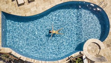 Vista aérea de una piscina con una forma curva distintiva, rodeada por un deck de baldosas y una estructura circular de piedra.