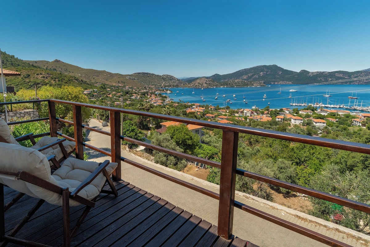 A balcony with wooden flooring and cushioned chairs, overlooking a marina with boats and a coastal town.