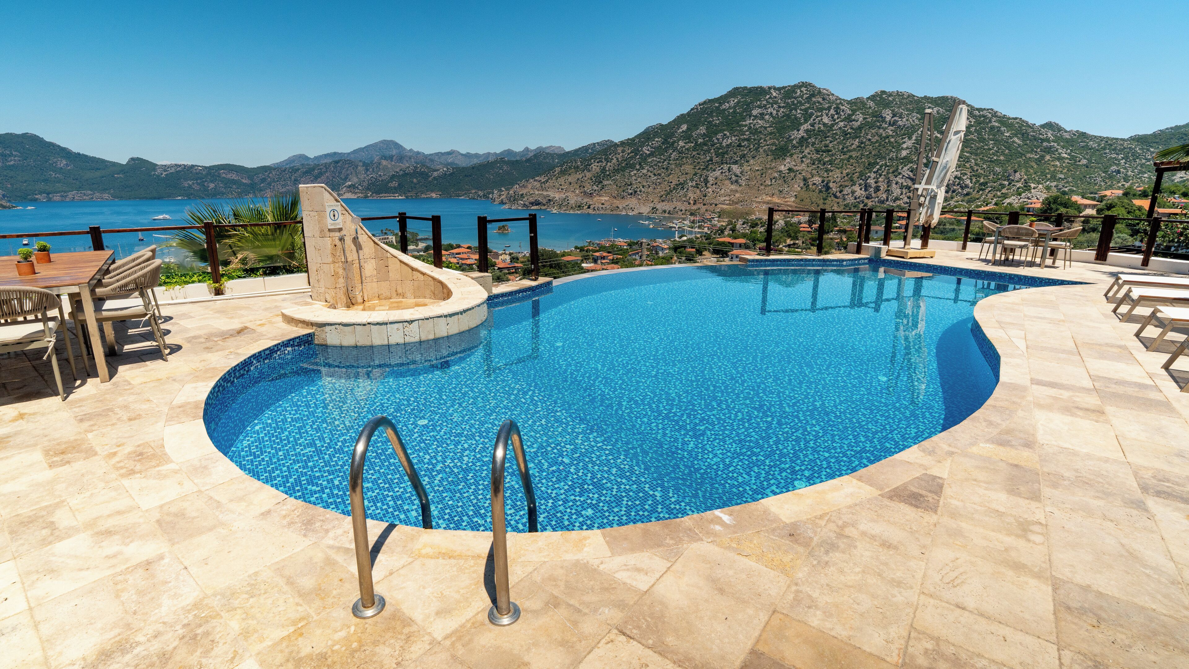 Une piscine avec vue sur la mer et les montagnes, entourée de mobilier d’extérieur et agrémentée d’une pergola.