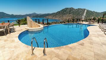 Ein Swimmingpool mit Blick auf das Meer und die Berge, umgeben von Gartenmöbeln und einer Pergola.