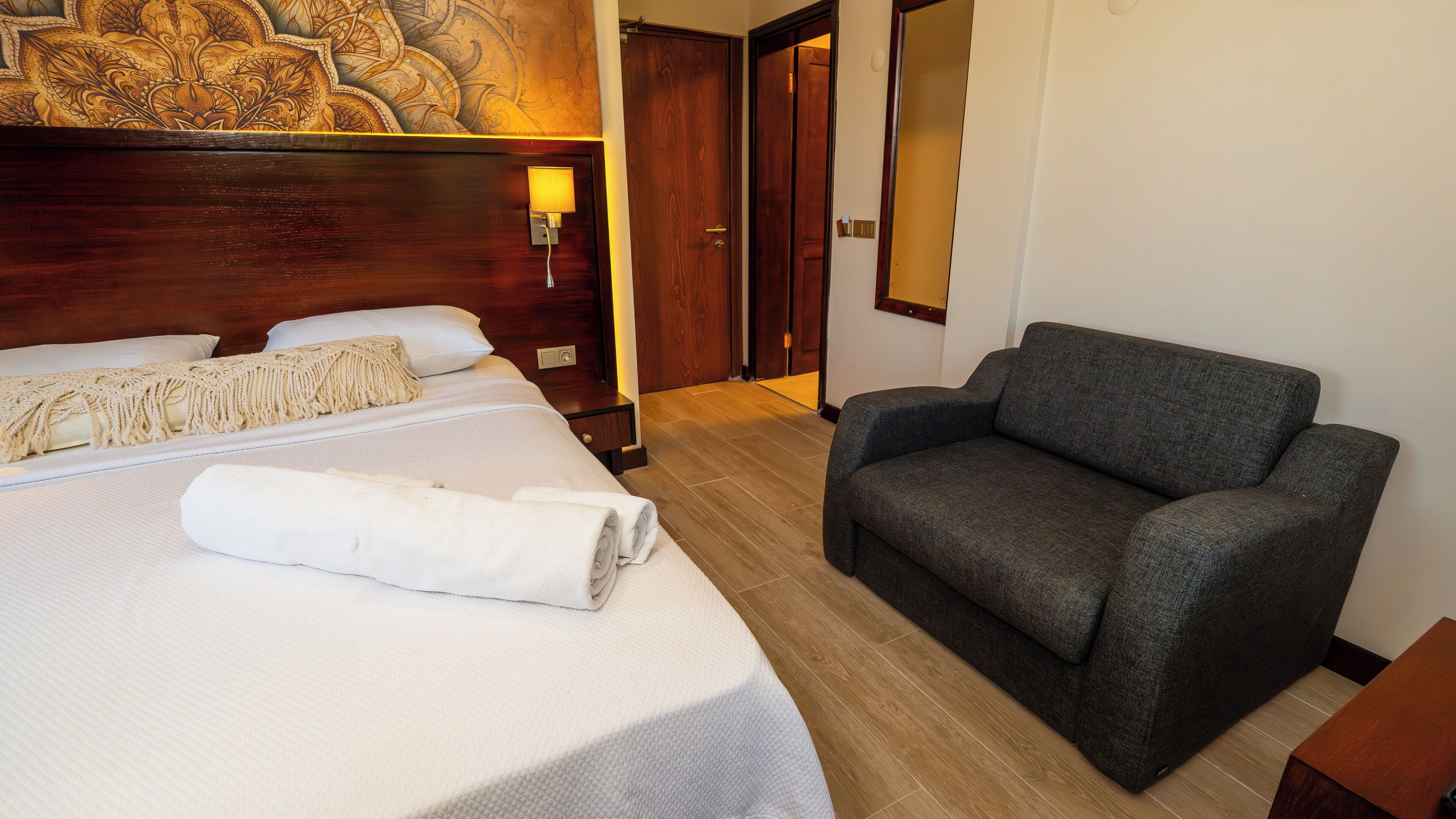A hotel room with a bed, a grey sofa, a wooden headboard, a wall-mounted lamp, and a door.