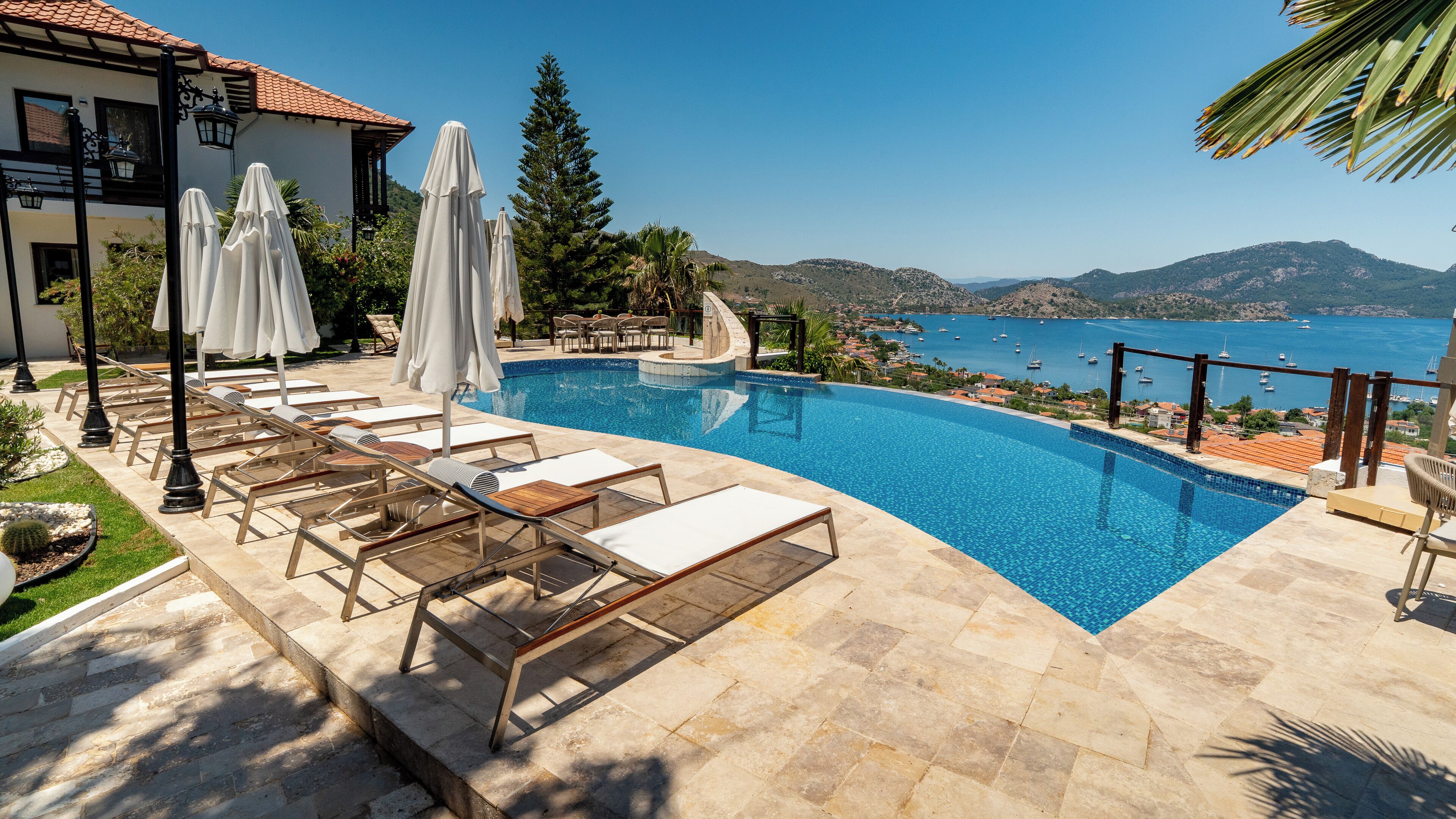 Zona de piscina con sillas de descanso y sombrillas, vista al mar y montañas al fondo.