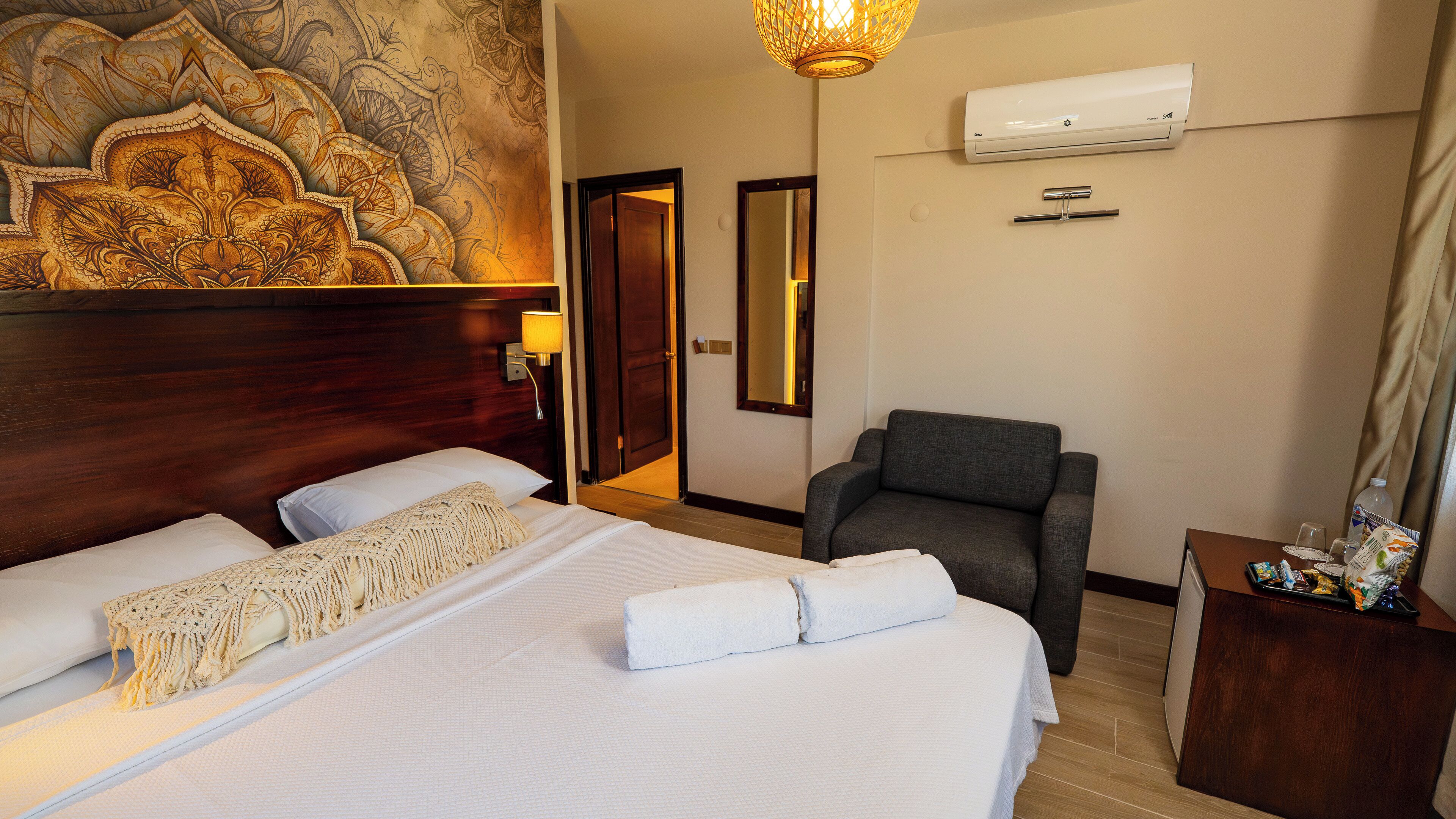 Ein Hotelzimmer mit einem großen Bett, einem Sessel, einem Schreibtisch mit Annehmlichkeiten, einer Wand-Klimaanlage und einem Wandbild.