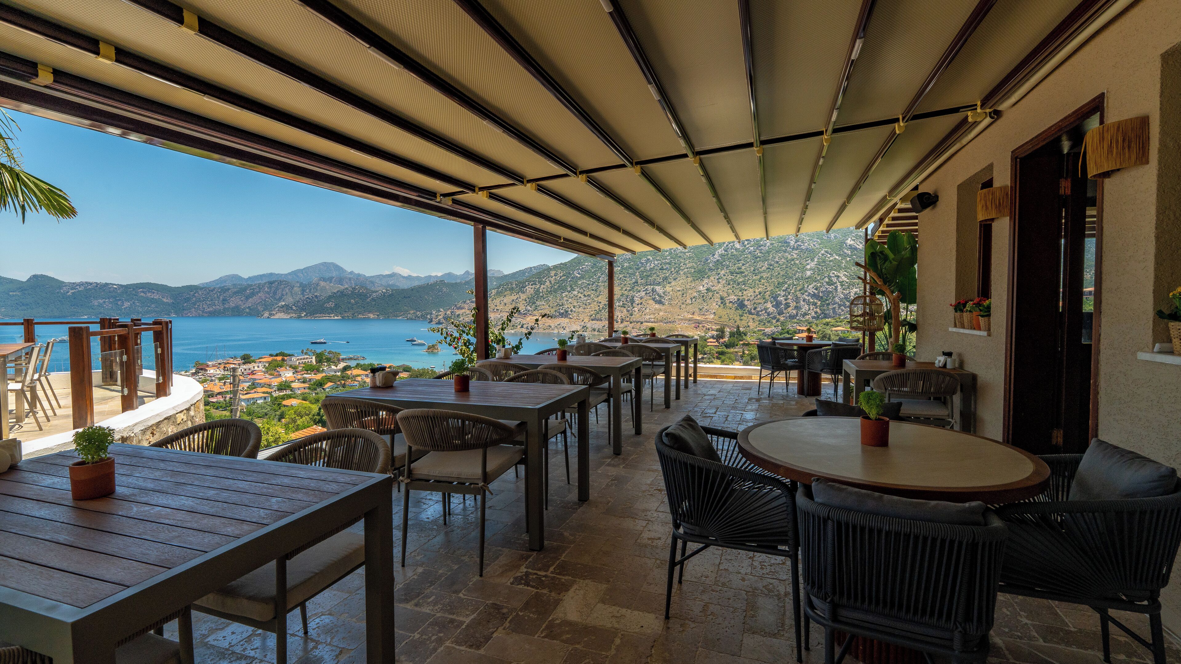 Terraza al aire libre con mesas y sillas, con vista al mar y montañas a lo lejos.