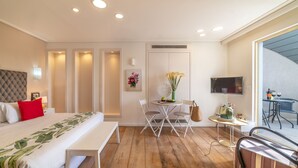 Una habitación de hotel moderna con una cama grande, una mesa de comedor con sillas, una mesita con un jarrón de flores y un televisor montado en la pared.