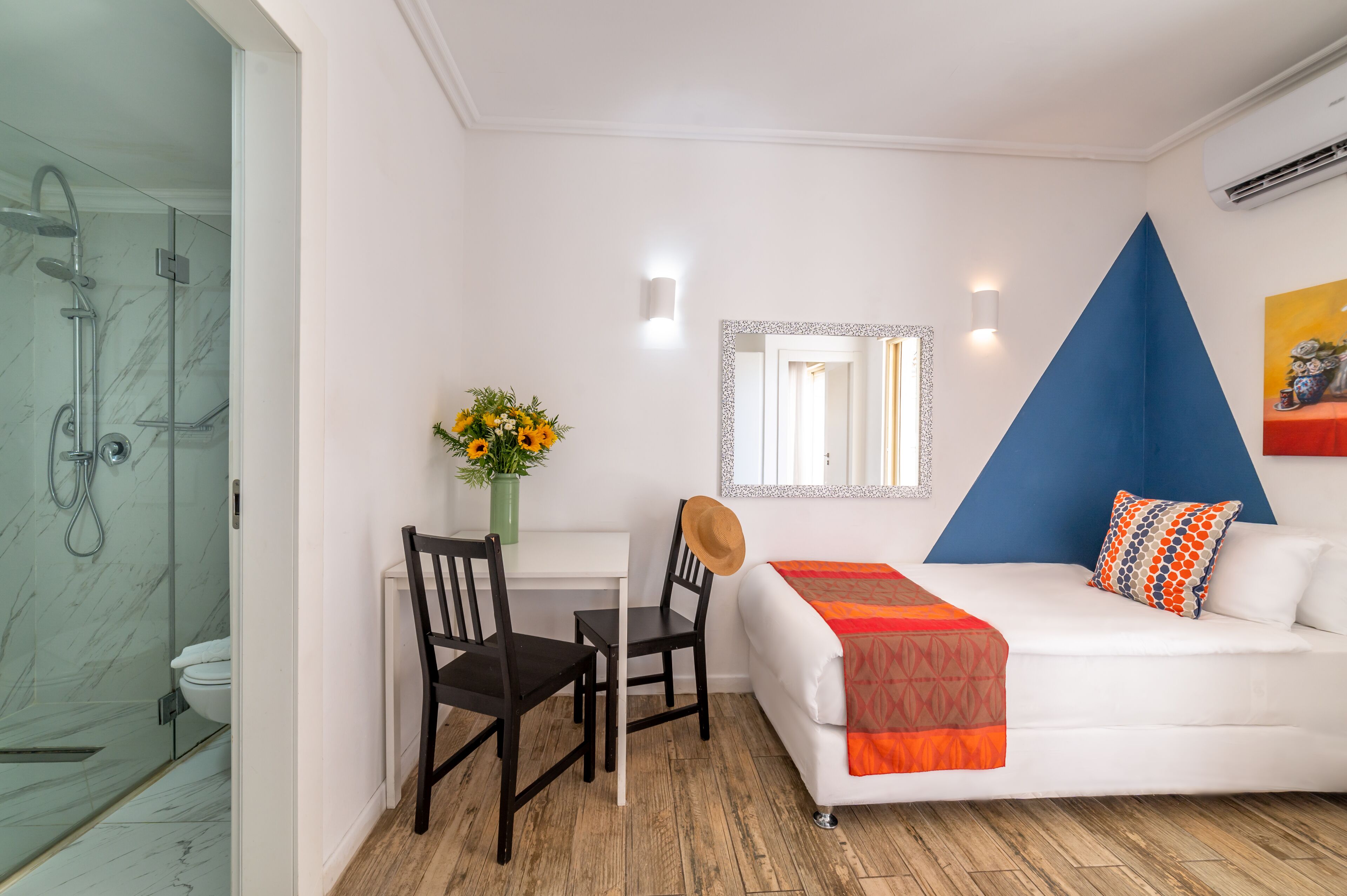 Una habitación de hotel moderna con ducha envuelta en vidrio, un sofá blanco con cojines naranja y azules, una silla de madera, una pequeña mesa con un jarrón de flores y un espejo con marco geométrico azul.