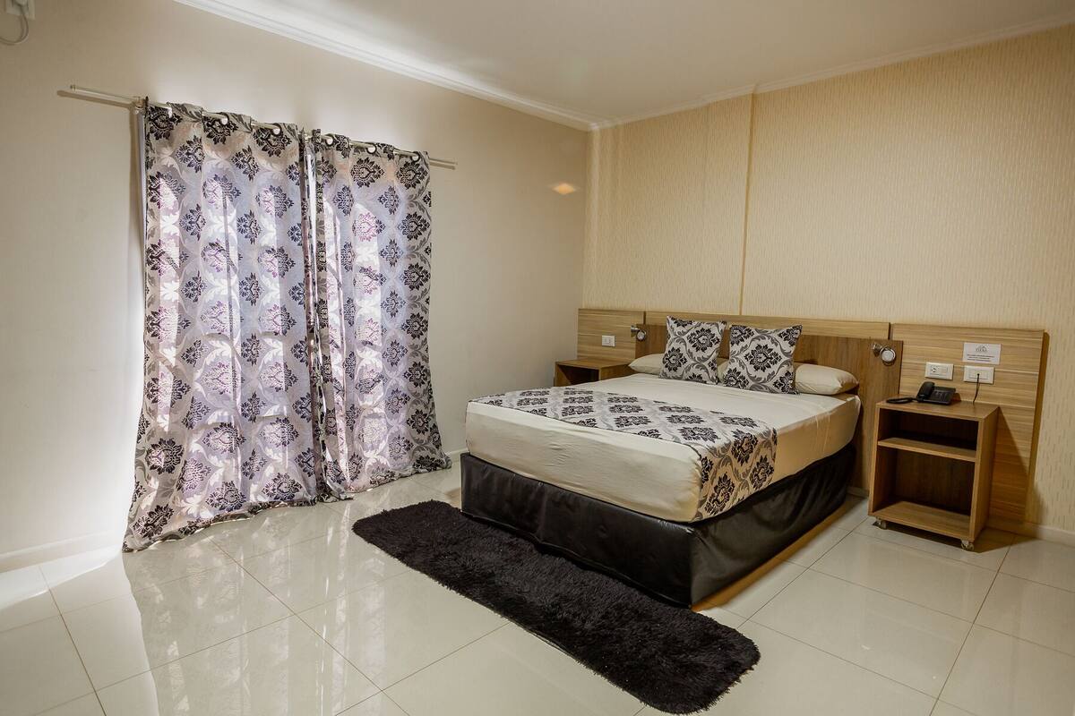 Un dormitorio con cama, mesita de noche, cabecero de madera, alfombra negra y cortina estampada con motivos florales.