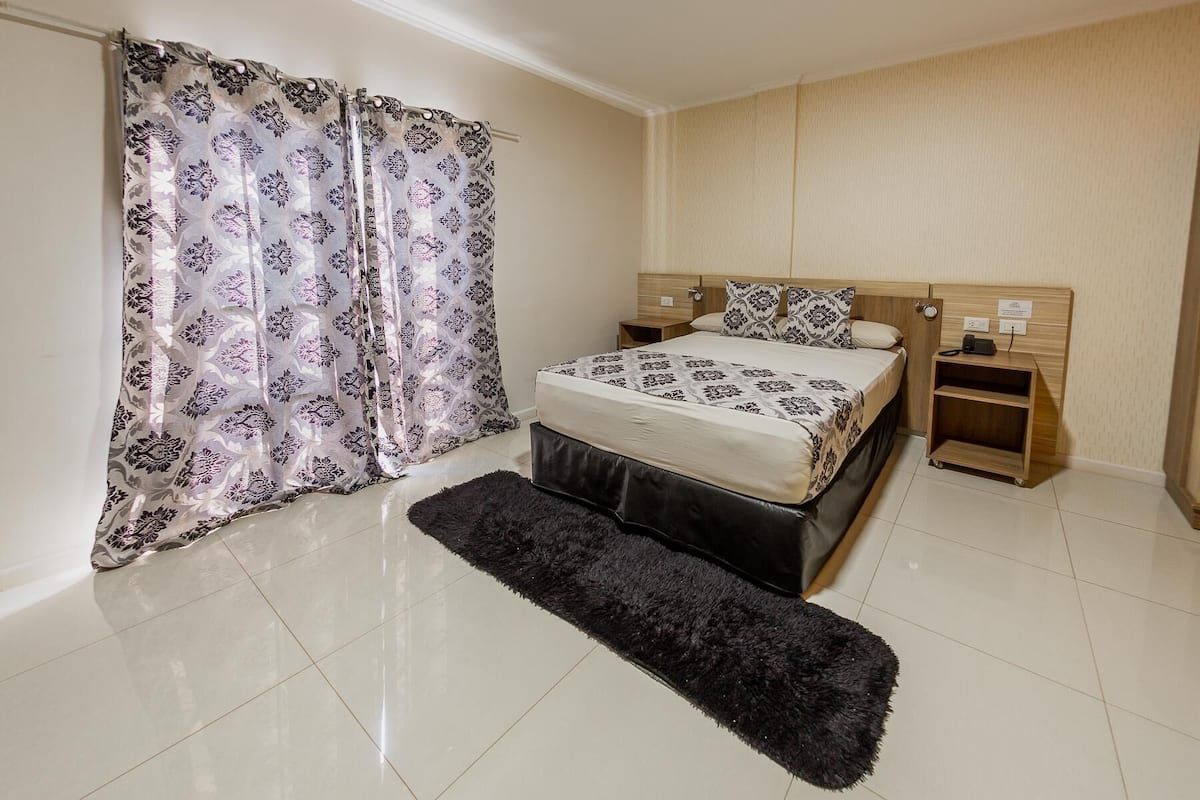 Un dormitorio con cama, mesita de noche, armario de madera, alfombra mullida negra y cortina estampada.