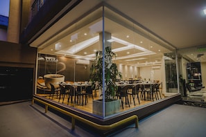 Een moderne hotellobby met een grote glazen wand, een centrale plant en een eetgedeelte met tafels en stoelen.