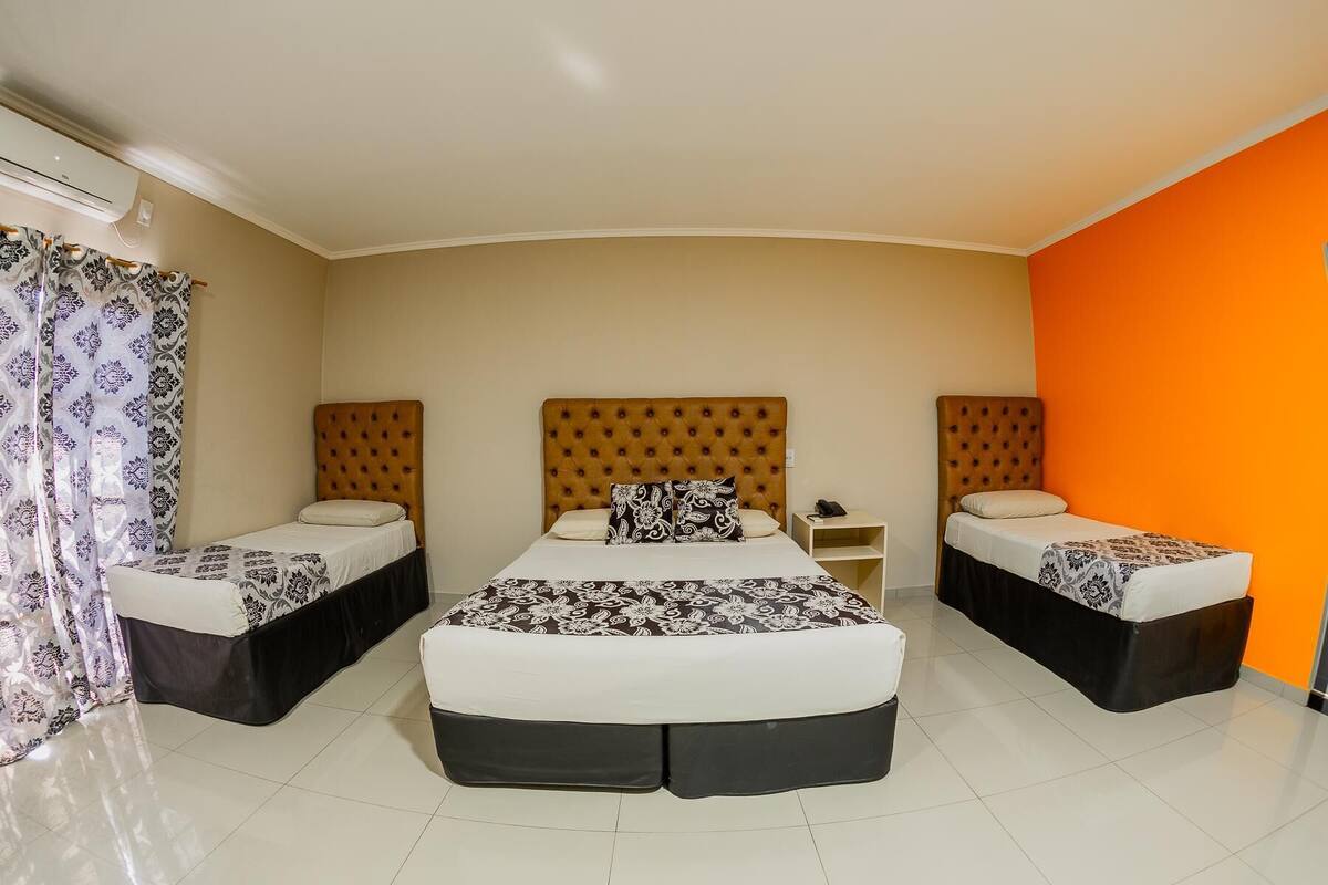 Habitación de hotel con una cama grande, dos camas más pequeñas, una mesita de noche y una pared de acento naranja.