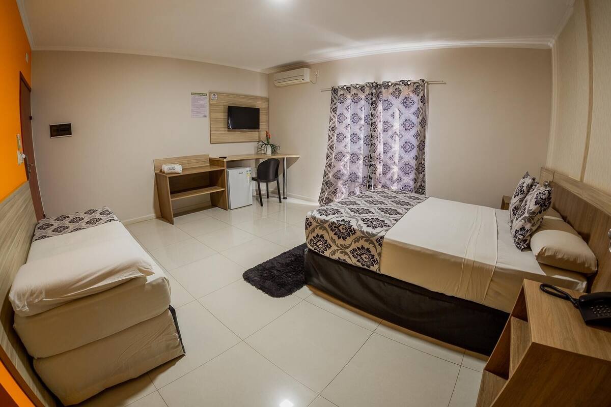 Habitación de hotel pequeña con cama, escritorio, silla, refrigerador compacto, televisión y cortina.