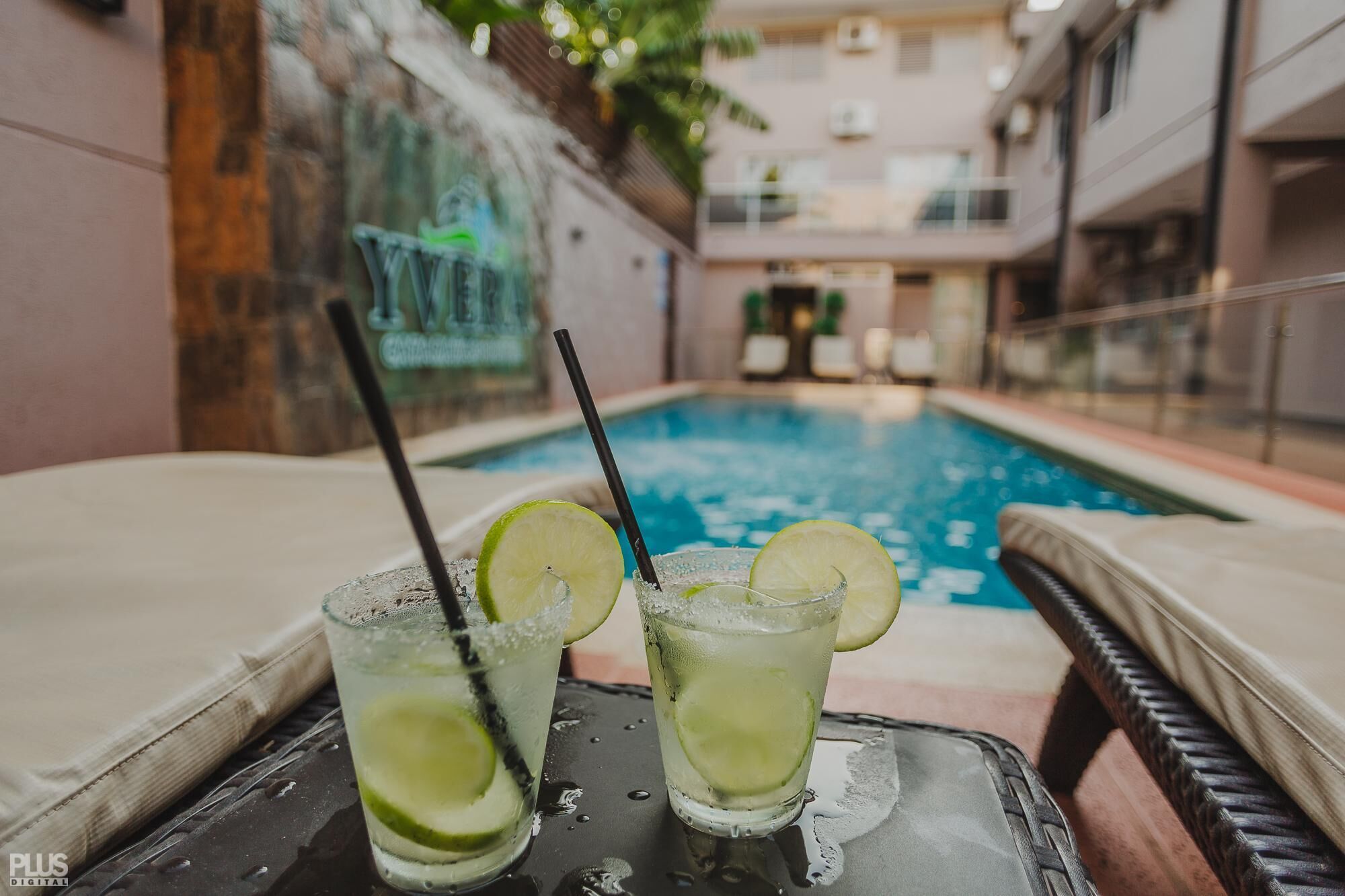 A poolside setting with two glasses of a lime-based beverage, a glass of water, and a black straw.