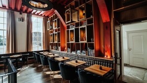 Una zona de bar con mesas de madera y sillas tapizadas de color negro, una estantería con diversos objetos y un amplio ventanal con cortinas.