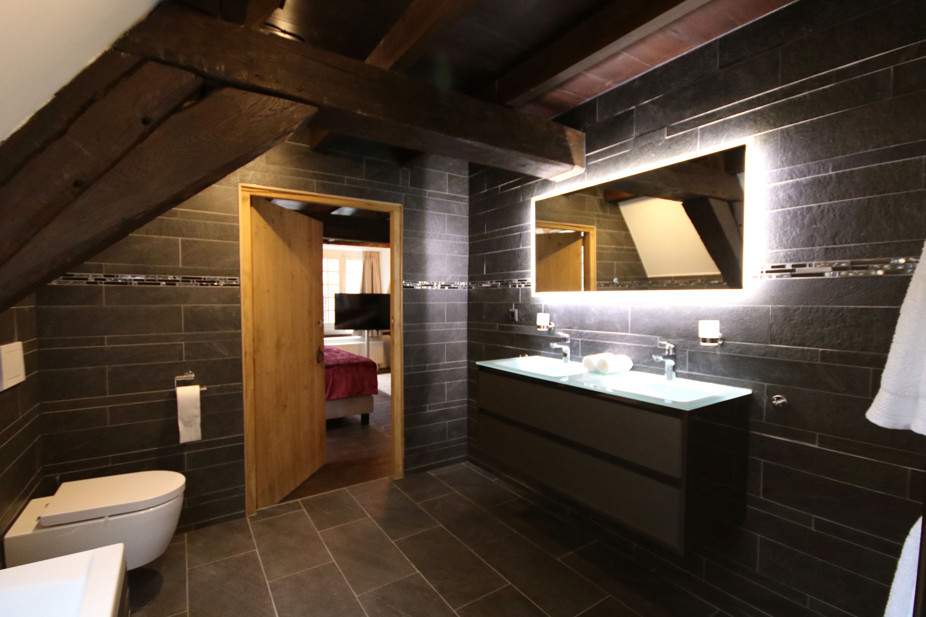 A modern bathroom with dark stone walls, a wooden ceiling, a white toilet, a sink with a mirror, and a door leading to a bedroom.