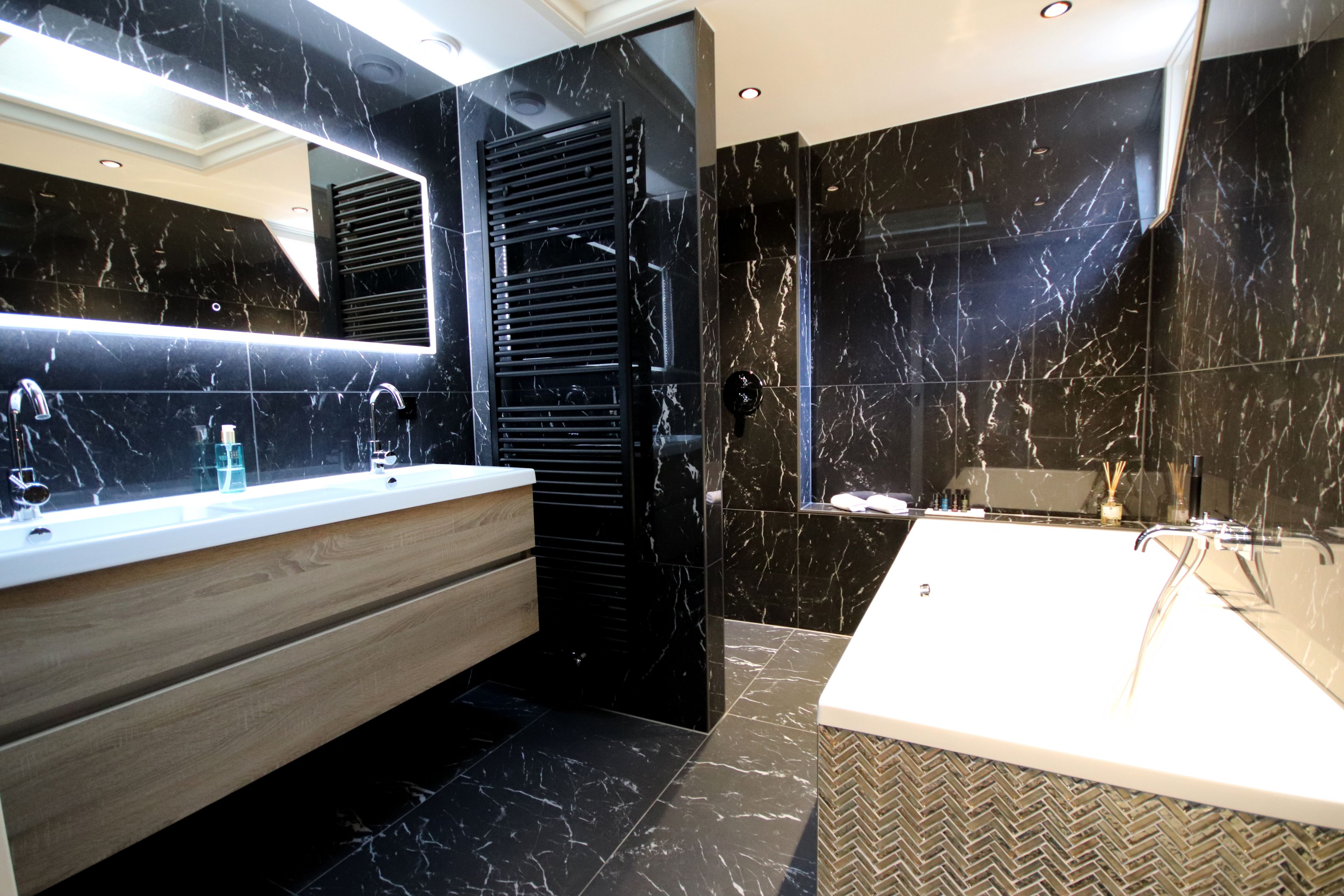 A modern bathroom with a large mirror, a wooden vanity, a bathtub, and black marble walls.