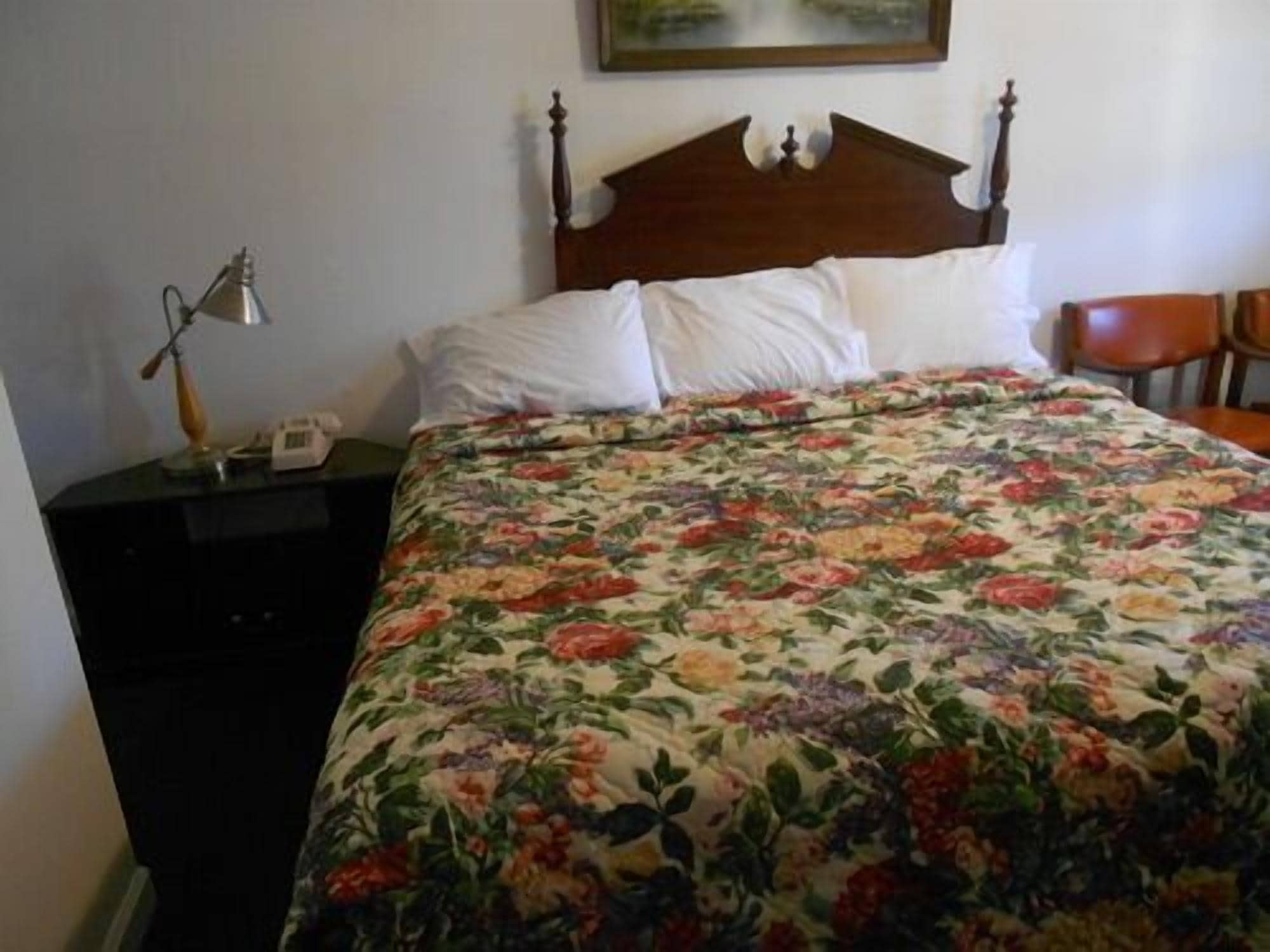A bedroom with a floral bedspread, a wooden headboard, a nightstand with a lamp, and a chair.