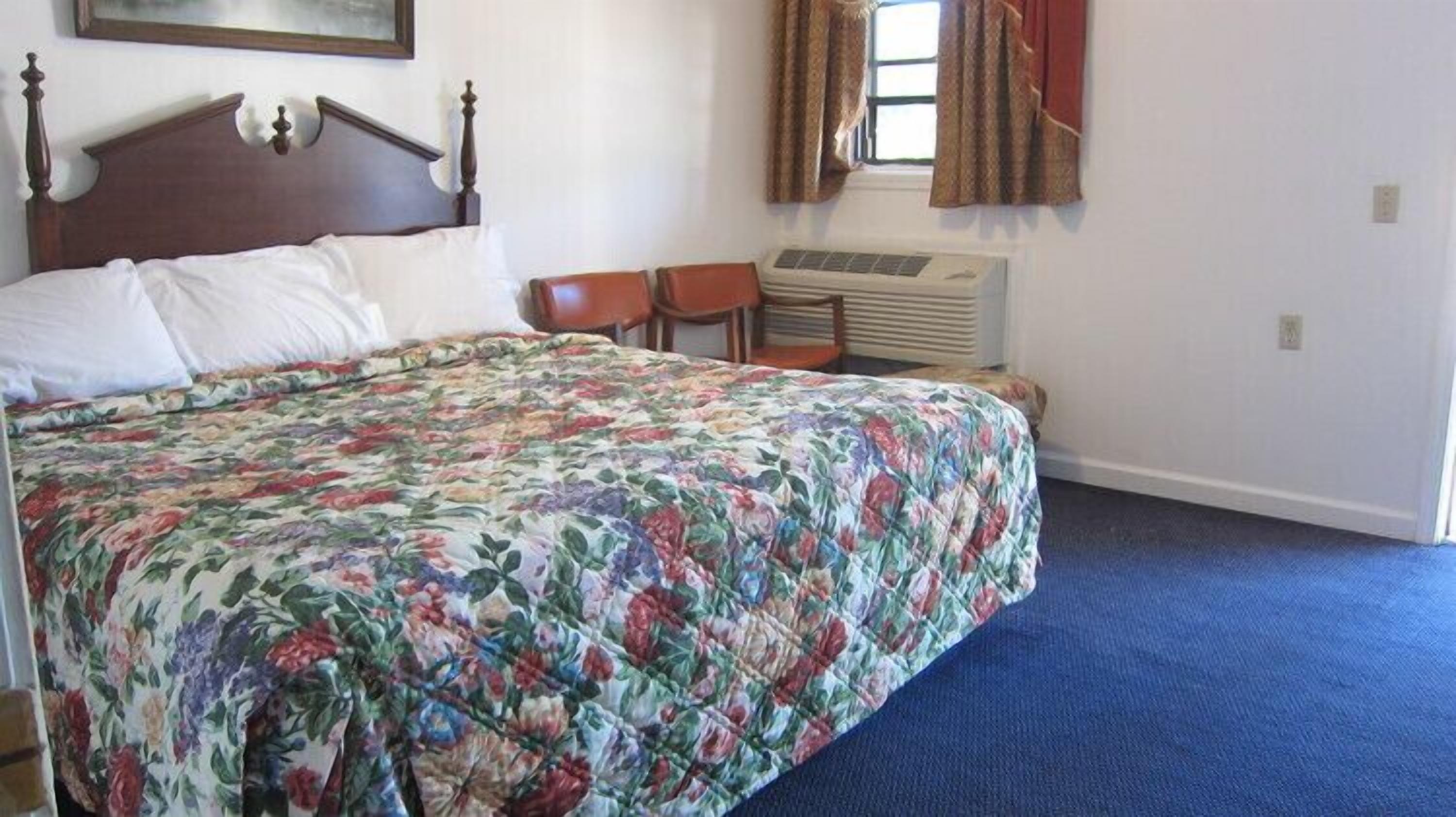 Una camera da letto con una coperta a fiori, una testiera in legno, due sedie, un condizionatore d'aria e una finestra con tende.