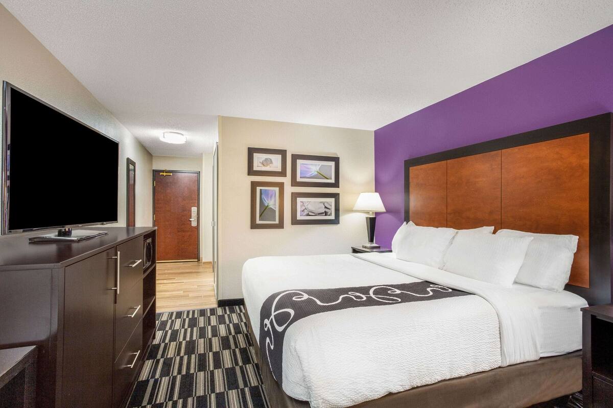 Ein Hotelzimmer mit einem großen Bett, einem Fernseher auf einer Holzkommode, einem Schreibtisch mit einer Lampe und einer Tür zu einem weiteren Raum.