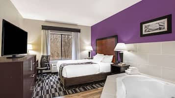 Habitación de hotel con cama, televisión, escritorio, silla, ventana con cortinas, baño con bañera e imagen colgada en la pared.
