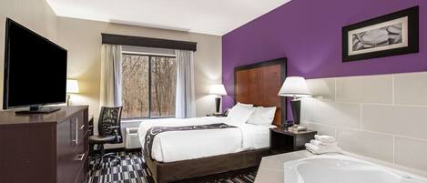 Habitación de hotel con cama, televisión, escritorio, silla, ventana con cortinas, baño con bañera e imagen colgada en la pared.