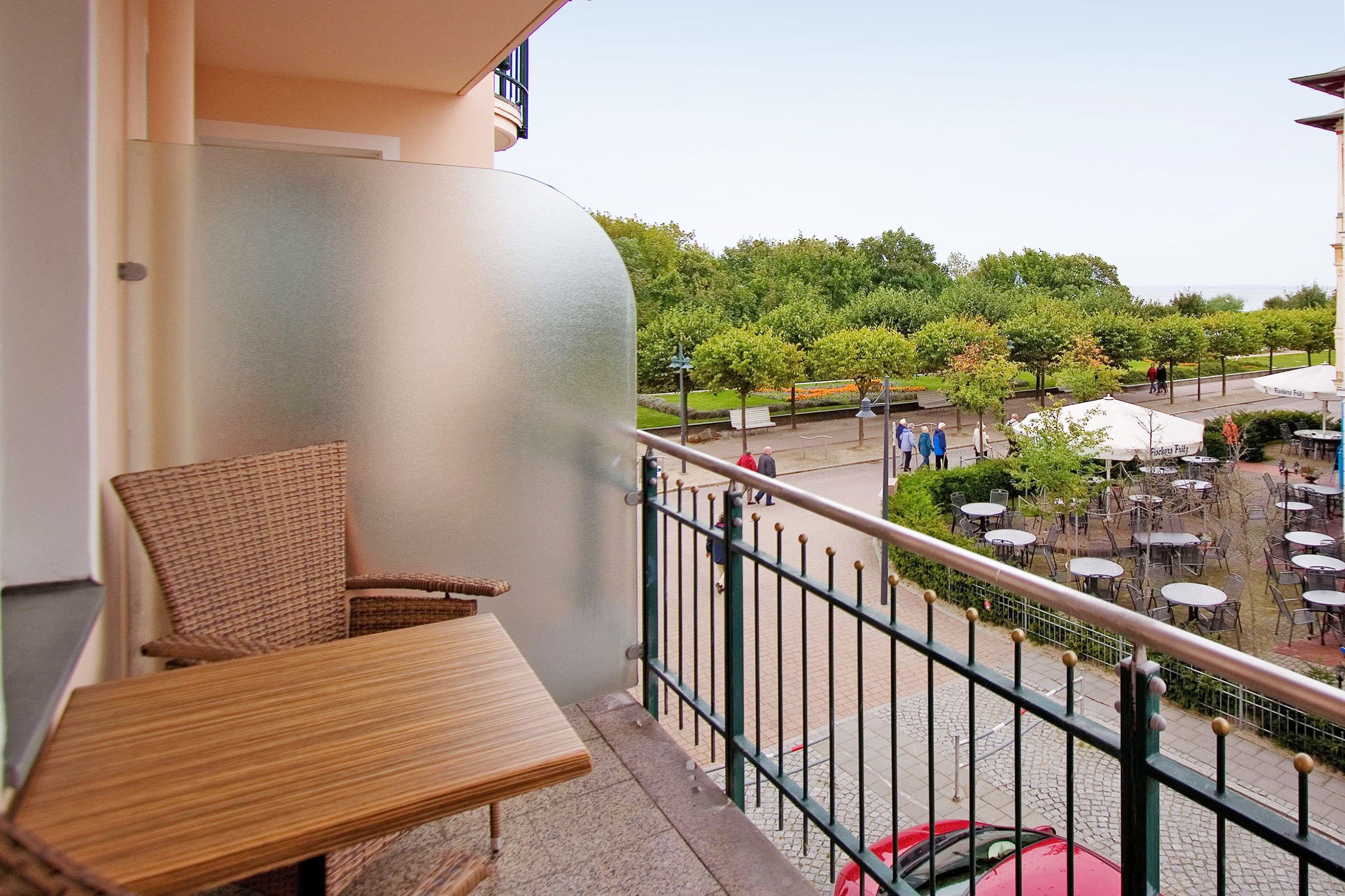 Un balcon avec des fauteuils en osier et une table, donnant sur une cour intérieure aménagée avec des sièges et où se trouve une voiture rouge.