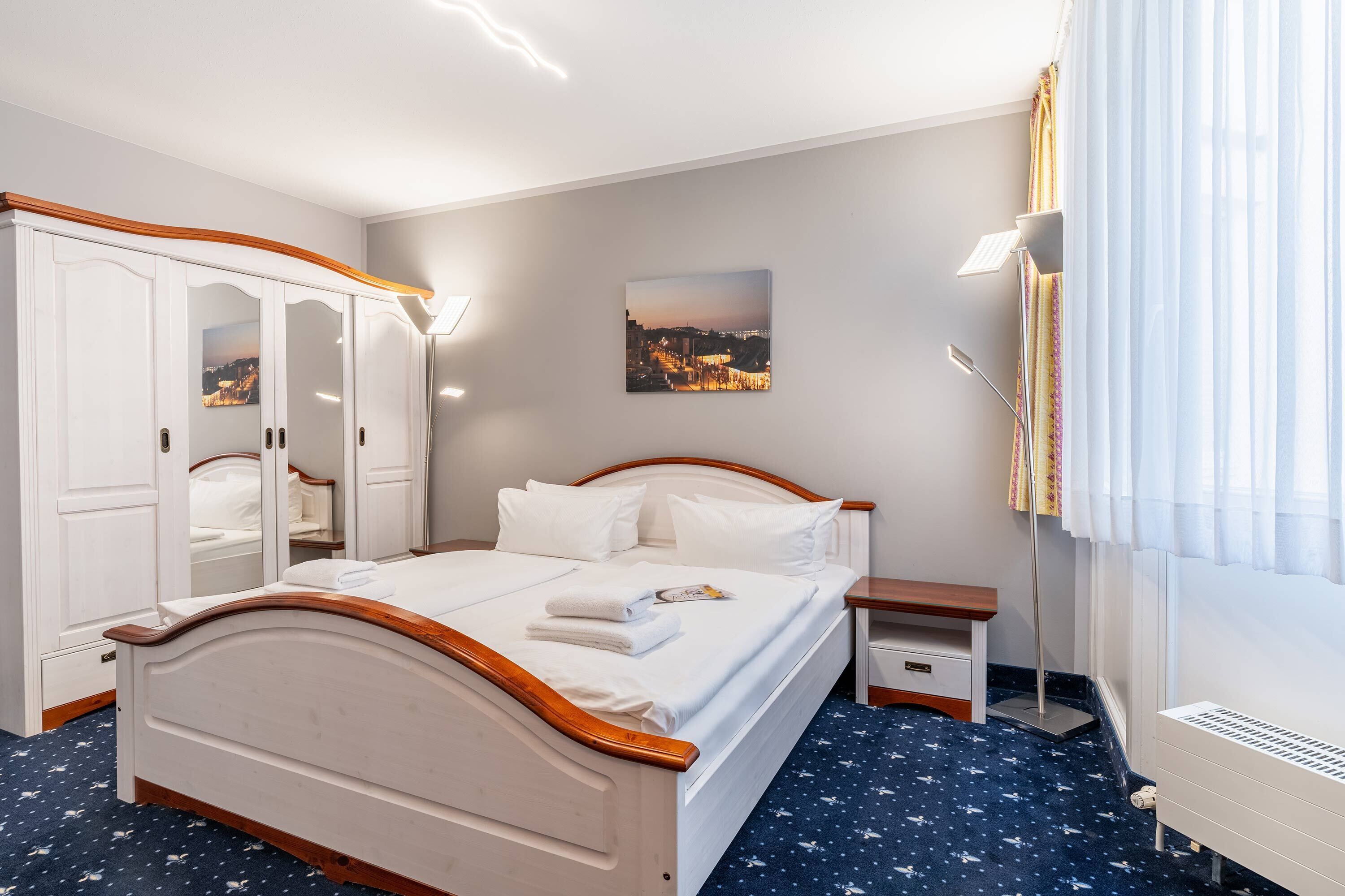 Une chambre d’hôtel avec un lit, une armoire, une table de chevet et un tableau représentant un paysage urbain accroché au mur.