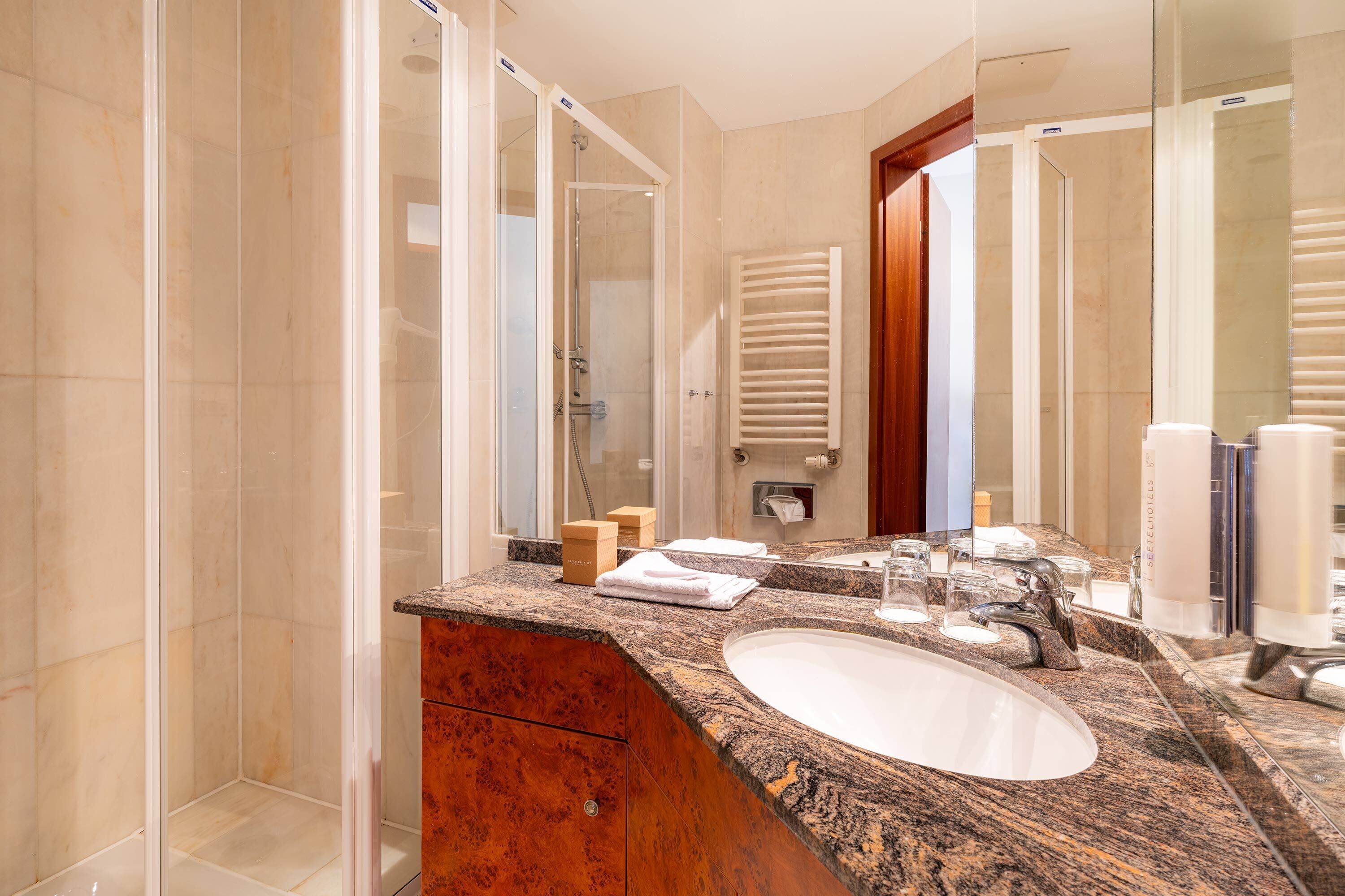A modern bathroom with a granite countertop, a white sink, a glass shower enclosure, and a wooden door.