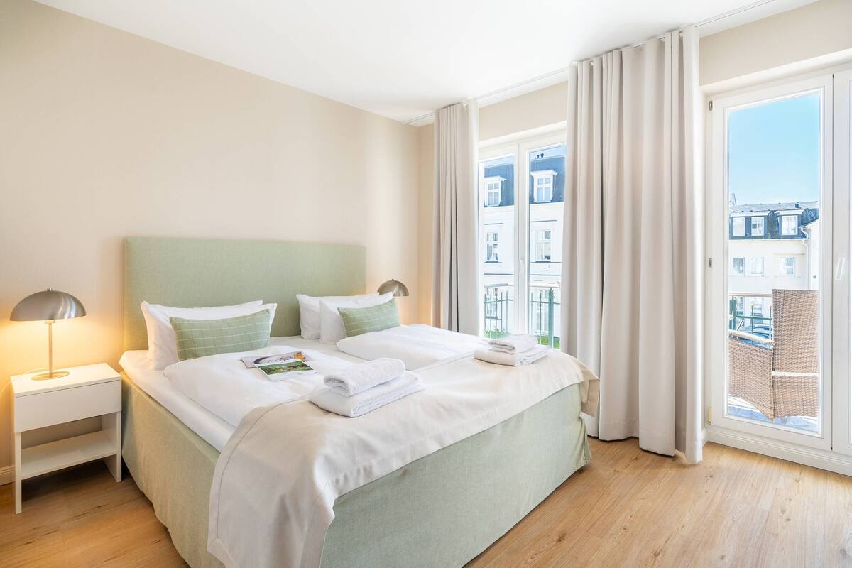 A hotel room with a large bed, a nightstand, a lamp, and a view of buildings through a window.