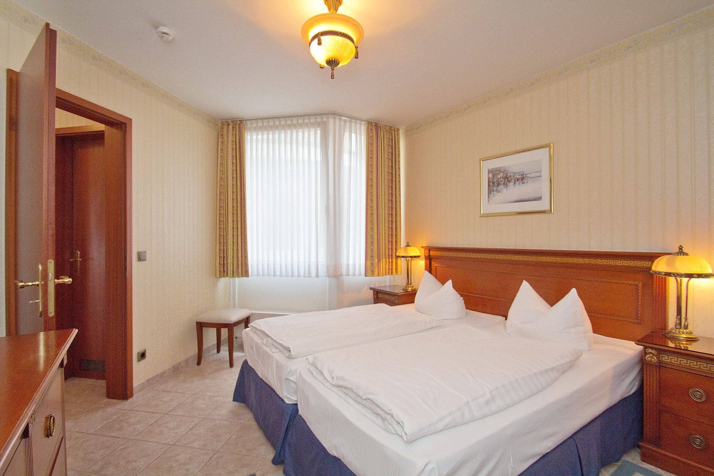 A hotel room with two beds, a desk, a chair, a nightstand with a lamp, a window with curtains, and a framed picture on the wall.
