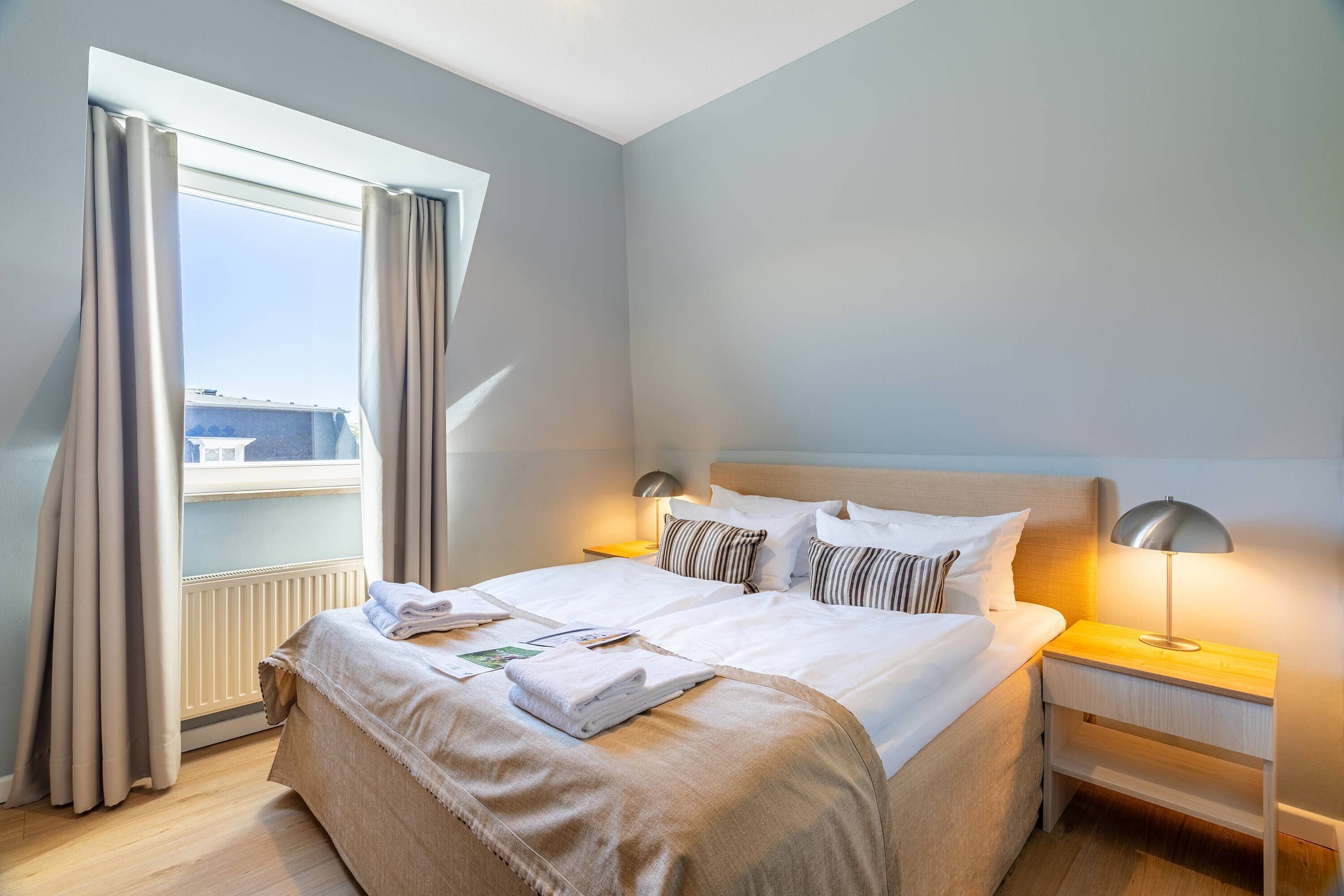 A neatly made bed with white linens, a bedside lamp, and a window with curtains.