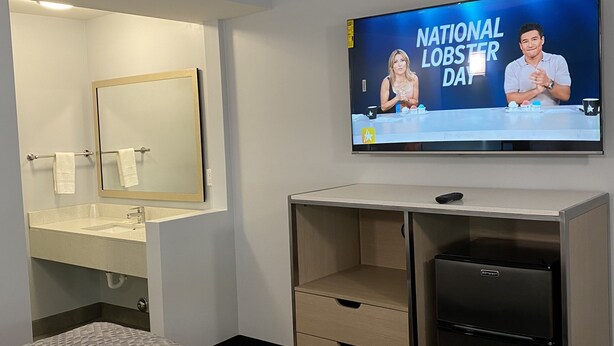 A hotel room with a bed, a TV displaying 'National Lobster Day', a mirror, and a minibar.