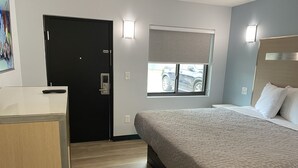 A hotel room with a bed, a desk, a door, a window with a view of a car, and wall-mounted lights.