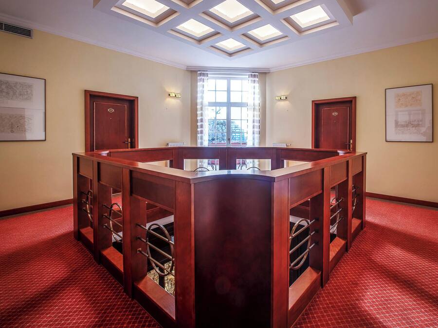 Quarto com escrivaninha de madeira central, duas portas, claraboia e piso de carpete vermelho.