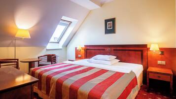 Una camera d'albergo con un letto, due comodini, una scrivania, una sedia, una lampada, una finestra e un quadro appeso al muro.