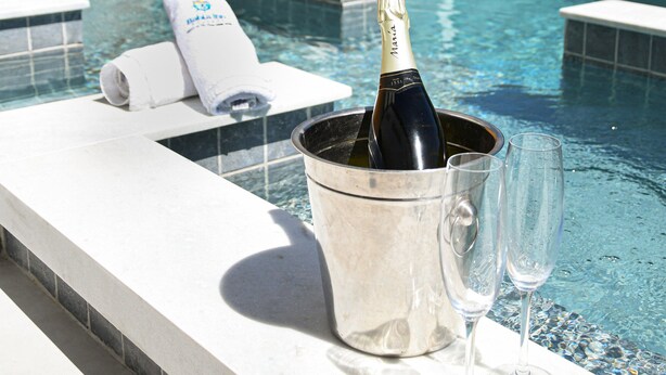 A poolside area with a stone wall, a sign, lounge chairs, a hot tub, and a bucket with a bottle and glasses. - Pousada Bahia Inn (Cairu)