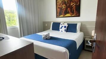 Quarto de hotel com cama, escrivaninha, um quadro na parede e janela com cortinas.