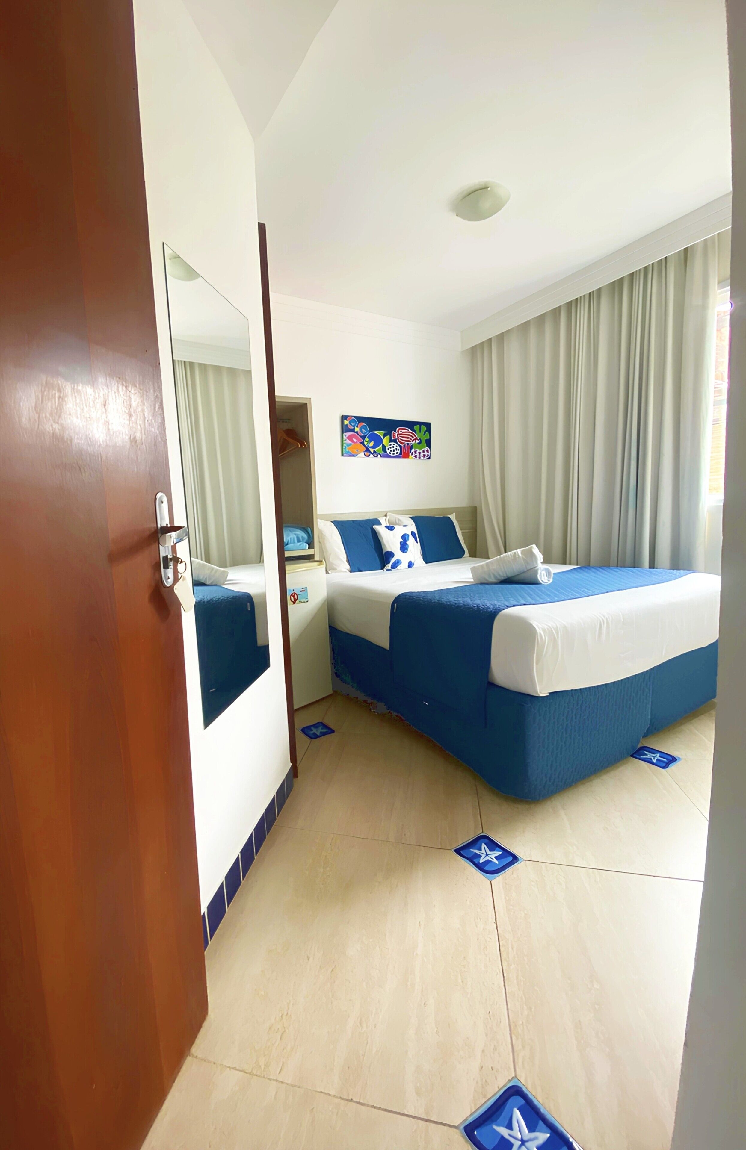 A hotel room with a bed, a bedside table, a mirror, and a window with curtains.