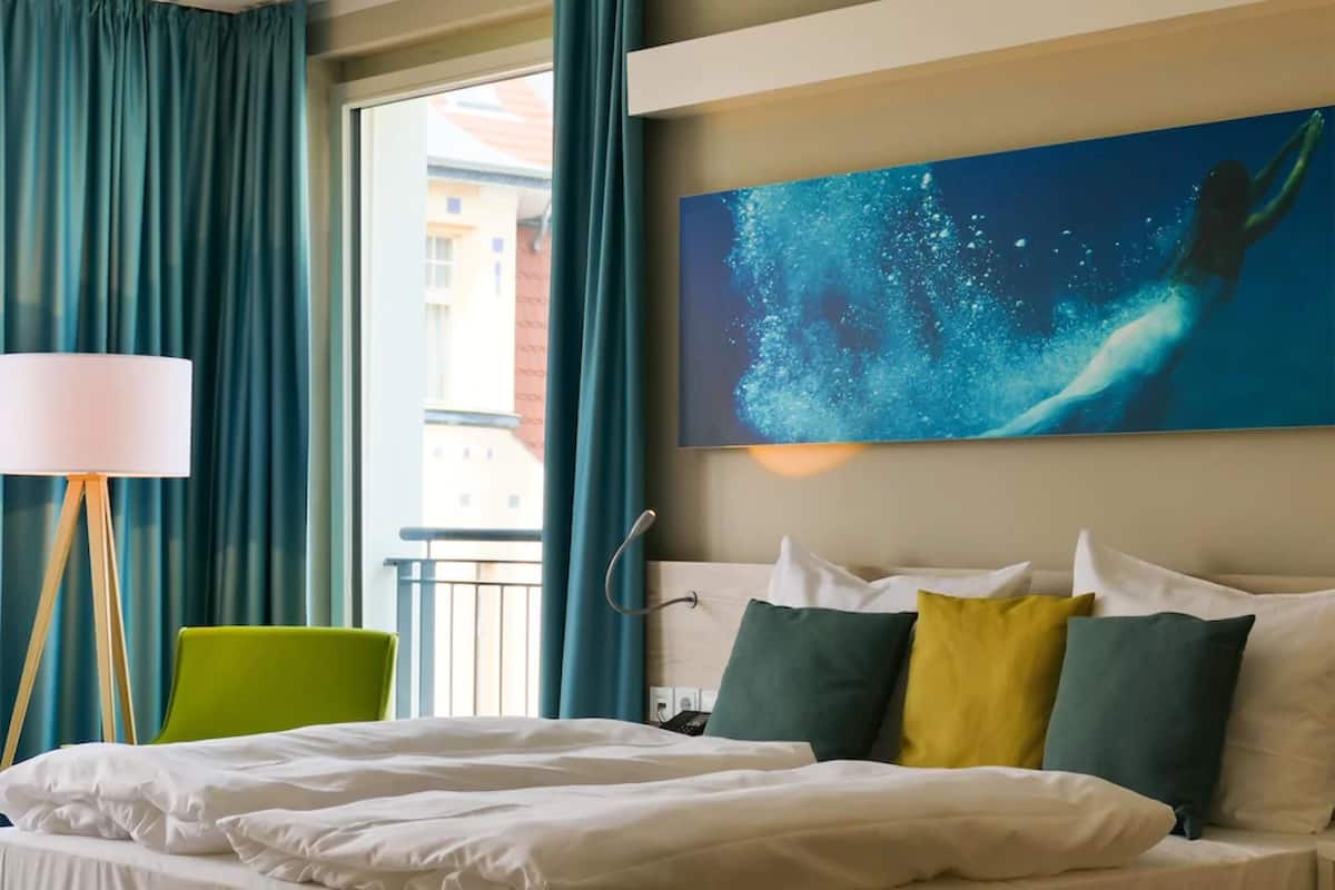 Habitación de hotel con cama, una silla verde, un cuadro de una sirena y vistas a edificios.