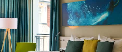 A hotel room with a bed, a green chair, a painting of a mermaid, and a view of buildings outside.