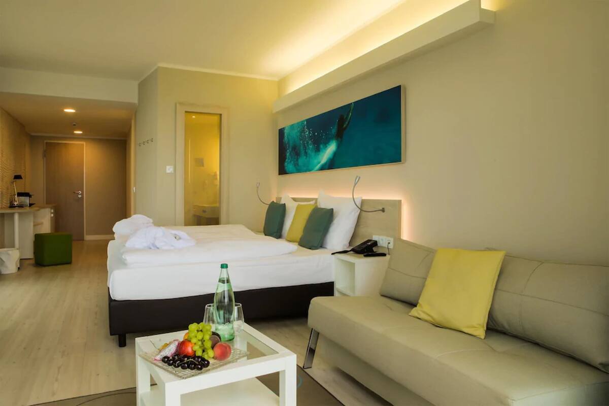 Una habitación de hotel con una cama, un sofá, una mesita de centro con fruta, un cuadro en la pared y un baño visible a través de una puerta abierta.