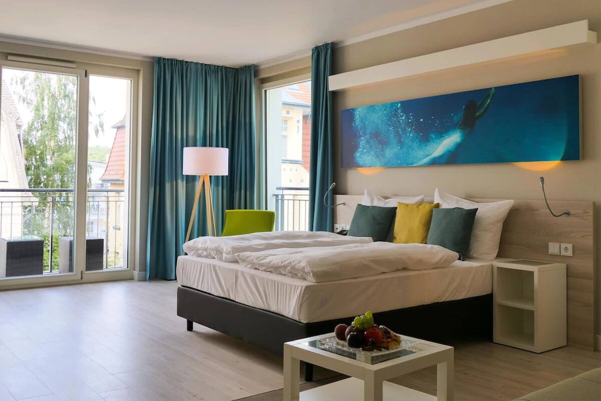 Una habitación de hotel moderna con una cama grande, vistas al exterior, un cuadro en la pared y una mesita con fruta.