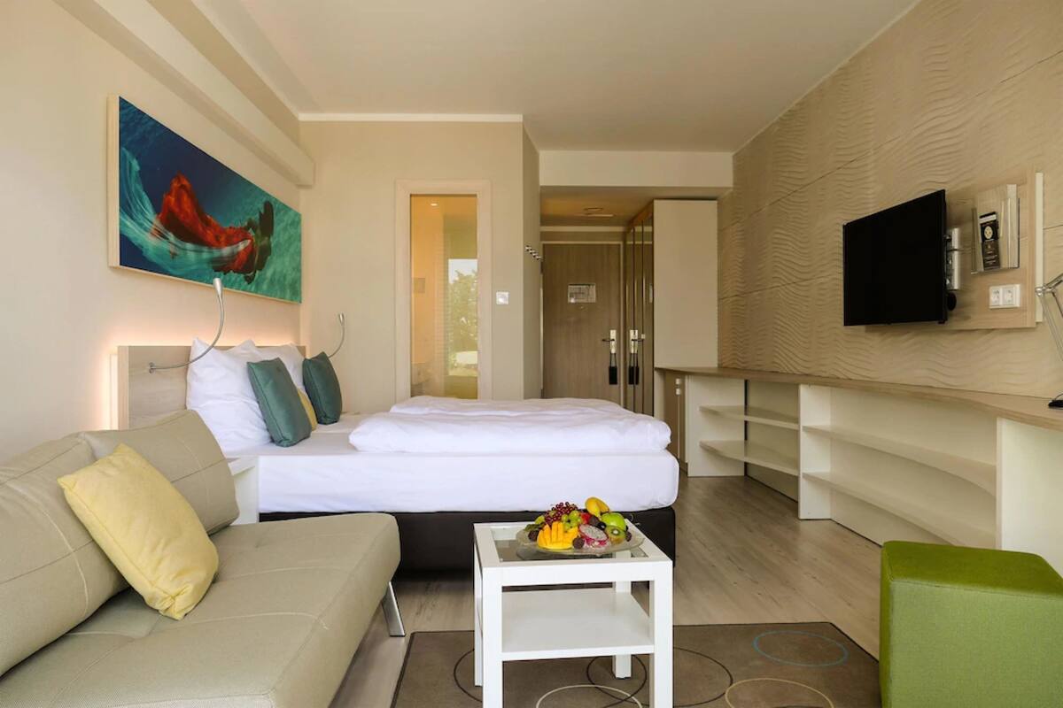 Habitación de hotel moderna con cama, sofá, una mesita con fruta, un cuadro en la pared y televisor montado.