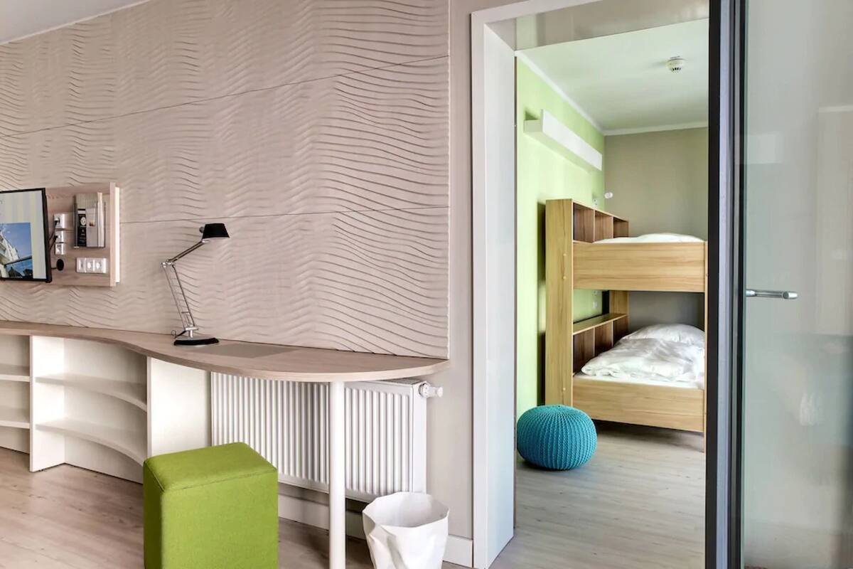 Un dormitorio moderno con una cama litera, un escritorio con lámpara, un radiador y un reposapiés verde.