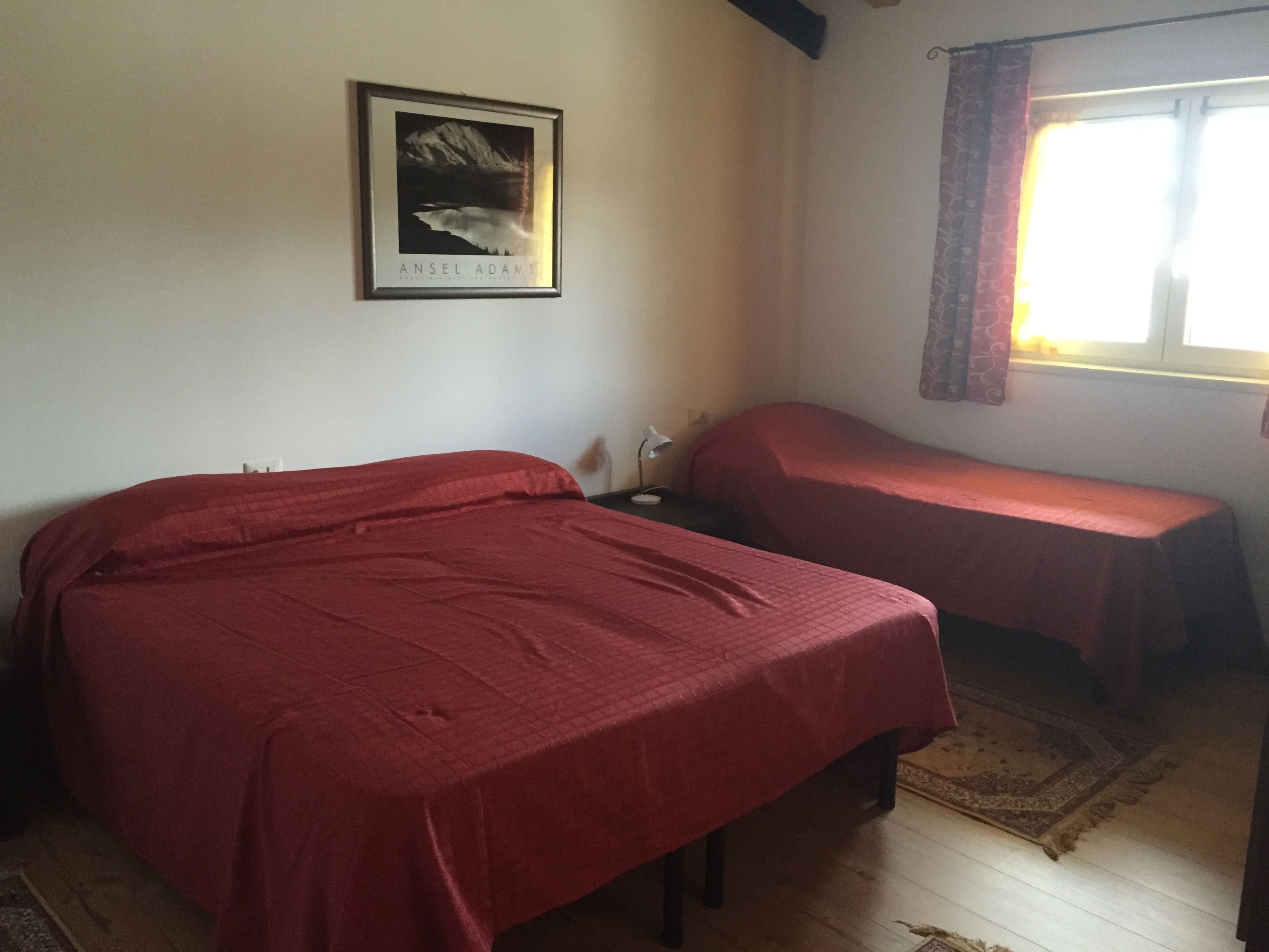 A room with two beds covered in red bedding, a framed picture on the wall, a bedside lamp, and a window with curtains.