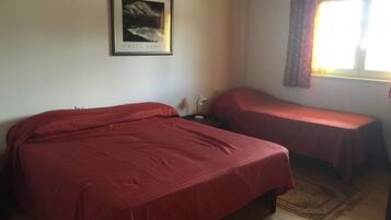 A room with two beds covered in red bedding, a framed picture on the wall, a bedside lamp, and a window with curtains.