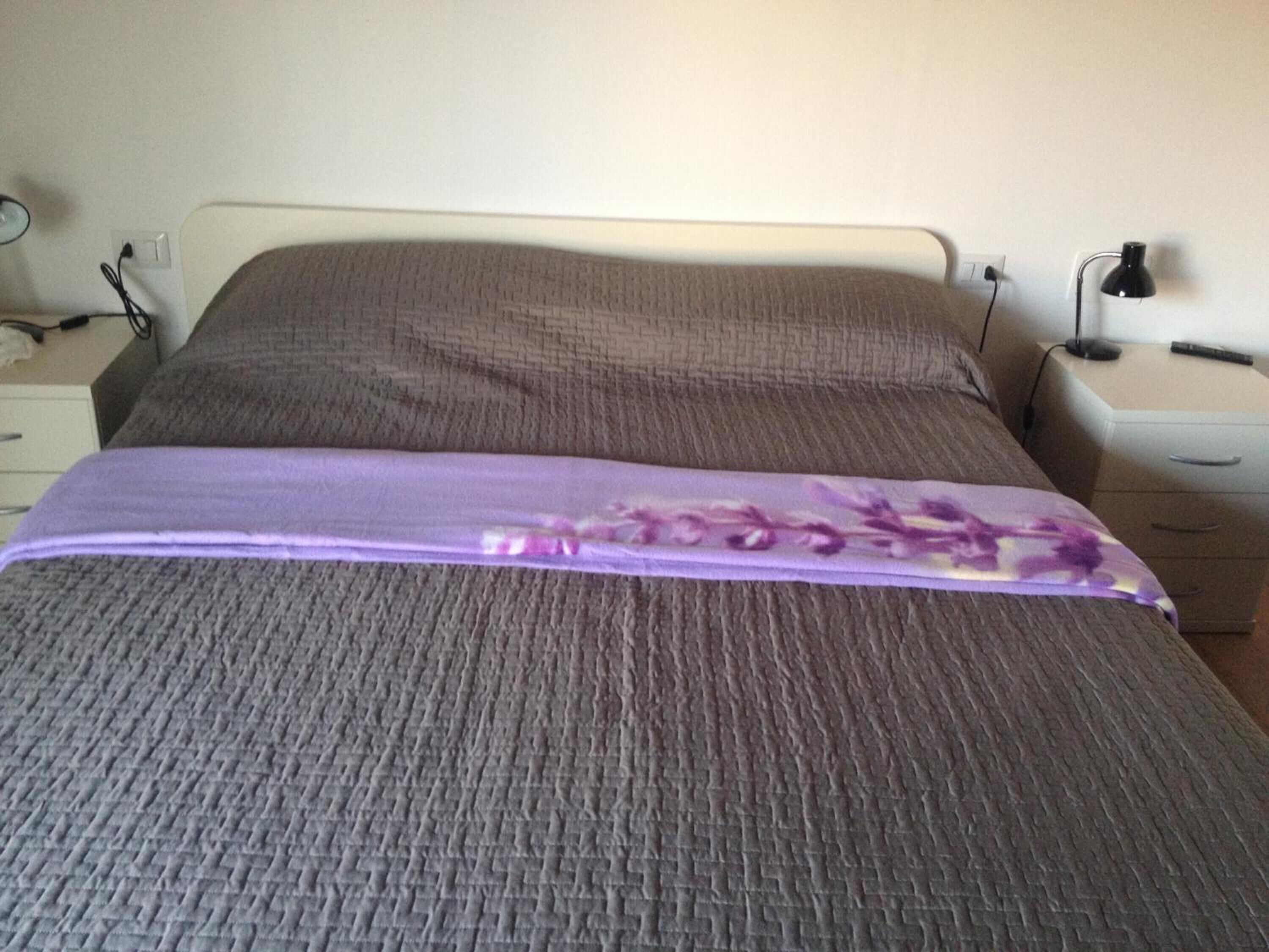 A bed with a grey textured bedspread and a purple floral blanket. There is a nightstand with a lamp and a power outlet on the wall.